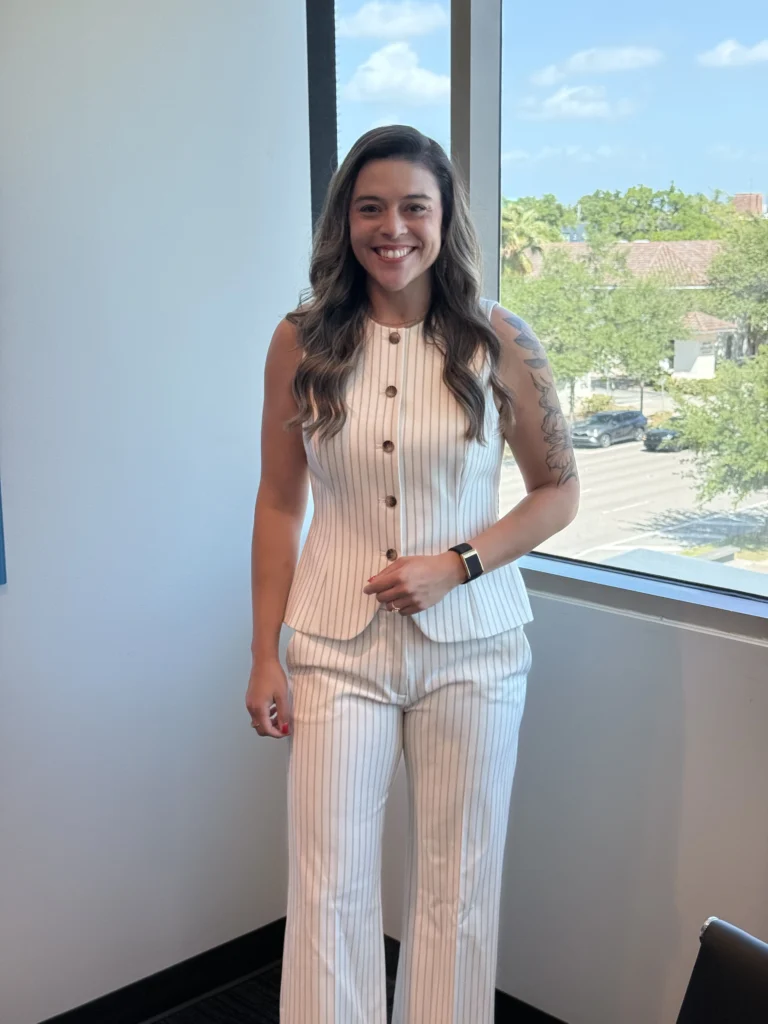 Nikki Savage standing in a bright office near a window wearing a white pinstriped outfit