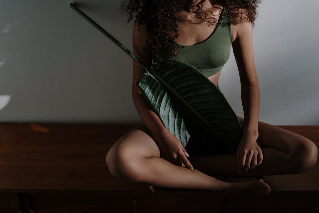 A serene photo of a woman meditating indoors, holding a large tropical leaf.
