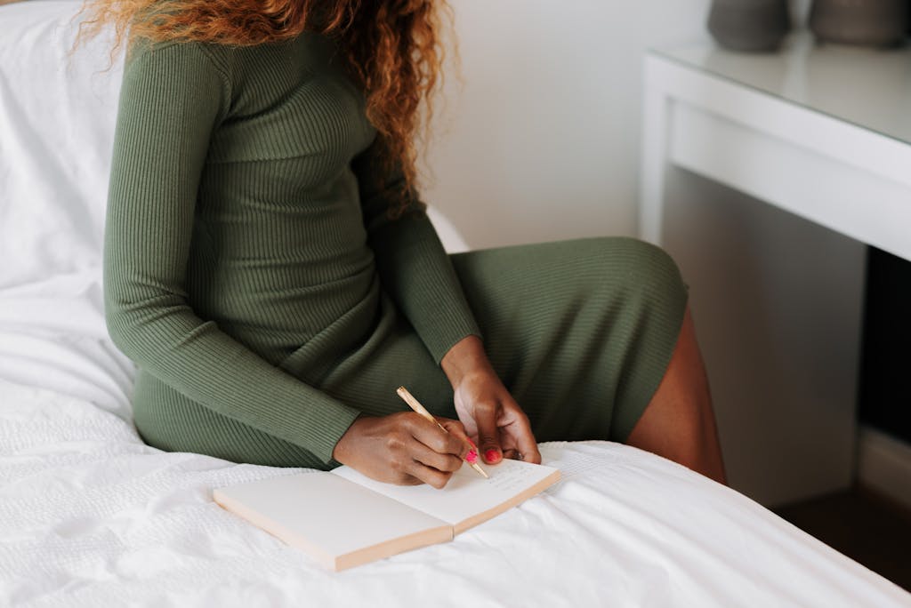 A woman in a green dress sits on a bed writing in a notebook, capturing a moment of calm and creativity.