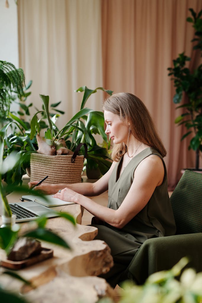 Businesswoman writing notes on laptop surrounded by lush indoor plants.