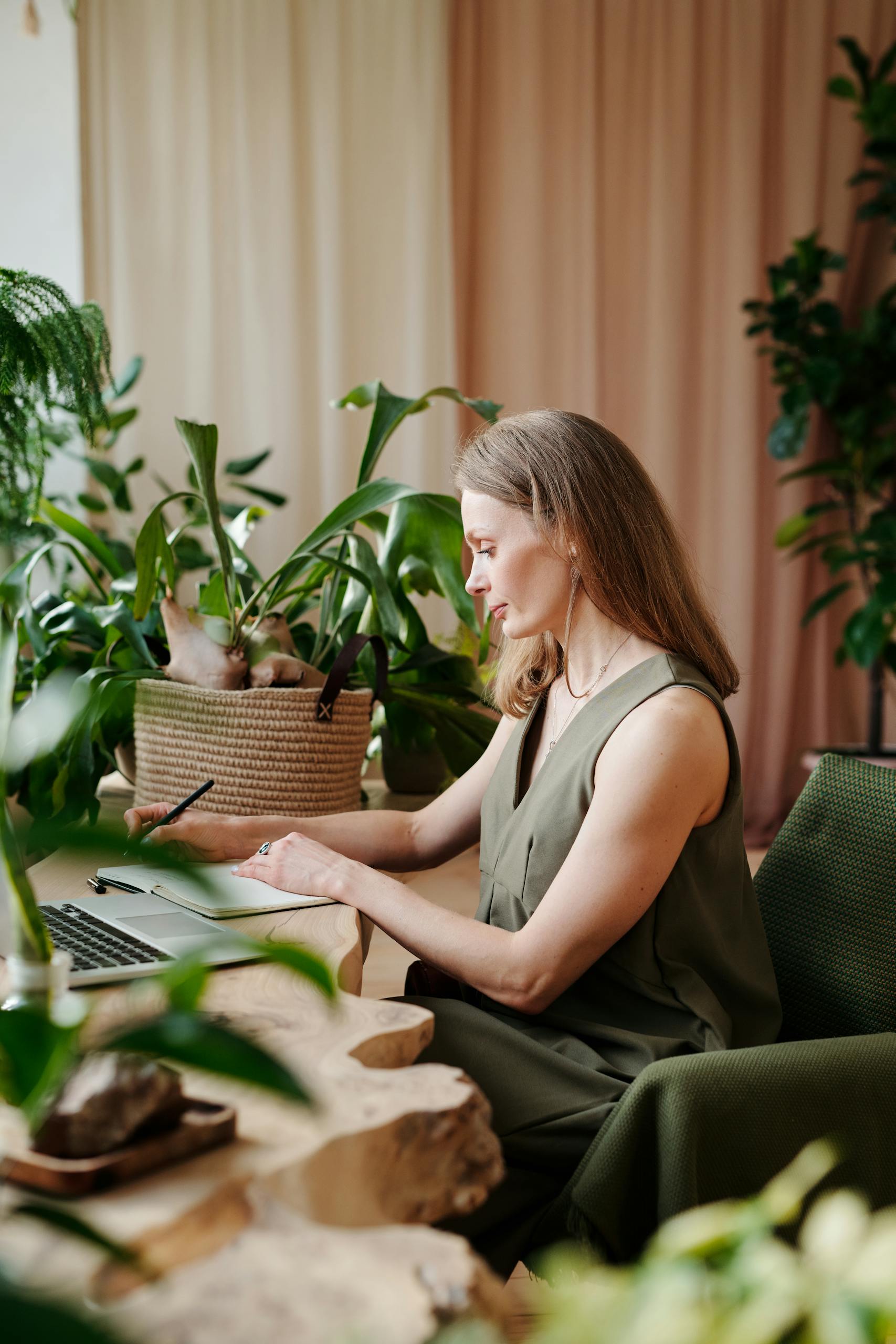 Businesswoman writing notes on laptop surrounded by lush indoor plants.