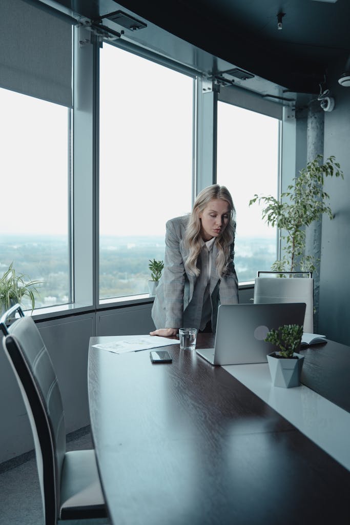 Focused businesswoman at laptop in modern office, large windows.